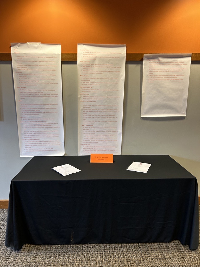 Three long scrolls of paper hung on the wall behind a table with a black tablecloth. On the table is a orange sign indicating the project (Drugs-Free Schools and Communities Act) and two printed reports laid on the table.