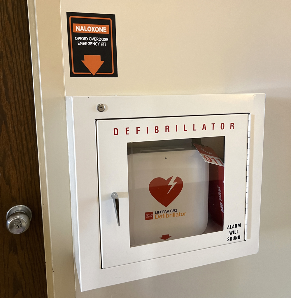 Orange and black sign with text stating "Naloxone Opioid Overdose Emergency Kit" above a defibrillator case on a white wall.