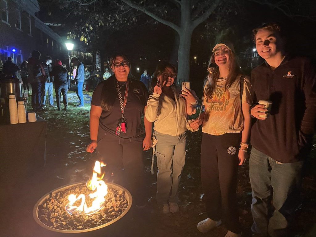 Individuals gathered around a fire roasting marshmallows with groups of people behind them faced the opposite direction.